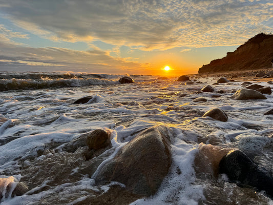 A scenic coastal sunset with waves washing over wet rocks in the foreground, a dark cliff on the right, and golden light reflecting off the water and clouds.