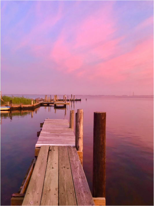 Weathered wooden dock extending into the calm Great South Bay beneath a pink and lavender sunset sky reflected in the water.