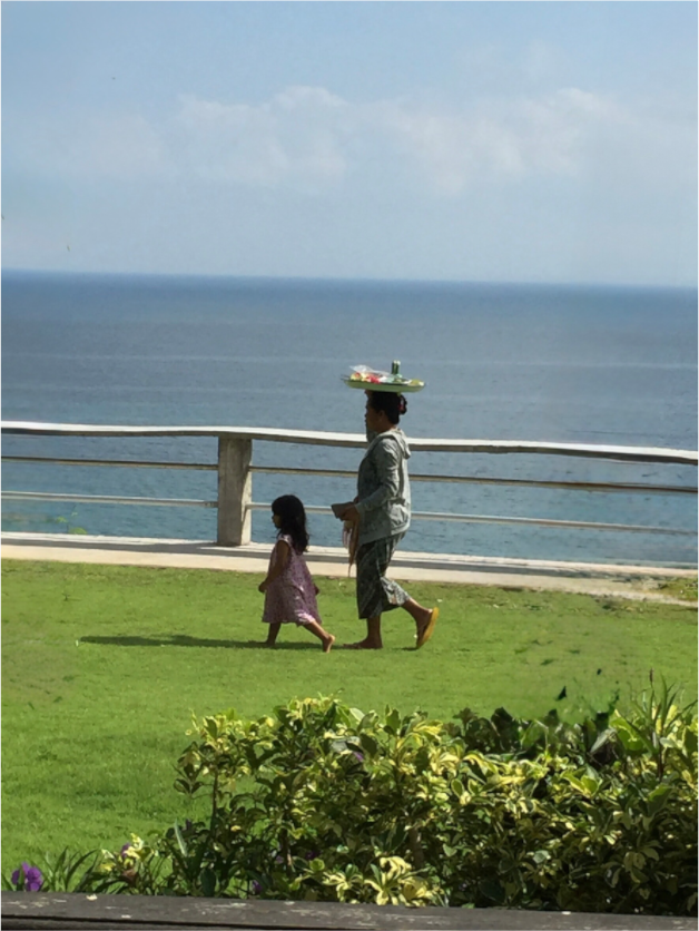Woman balancing a tray on her head while walking beside a young girl overlooking the Indian Ocean in Uluwatu, Bali, Indonesia.
